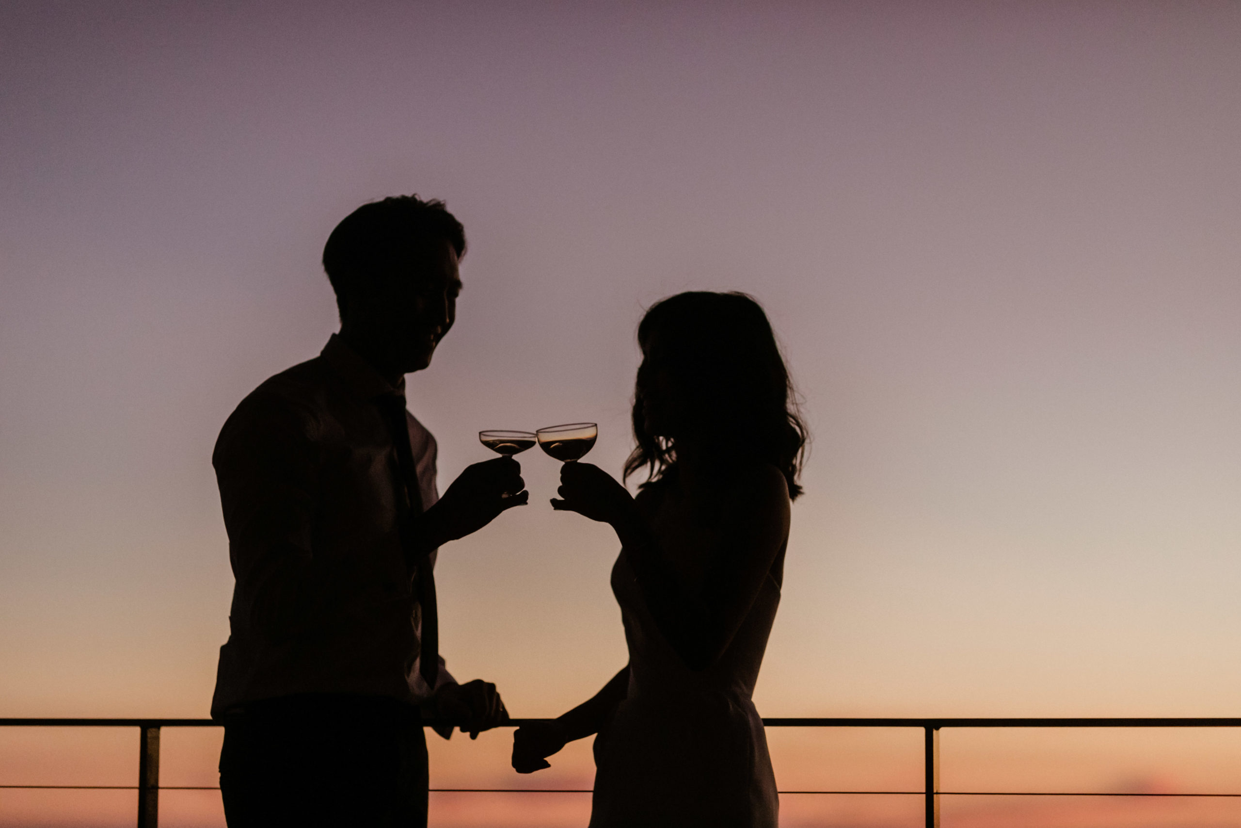 bride and groom pose together with their drinks in hand and sunset in the background