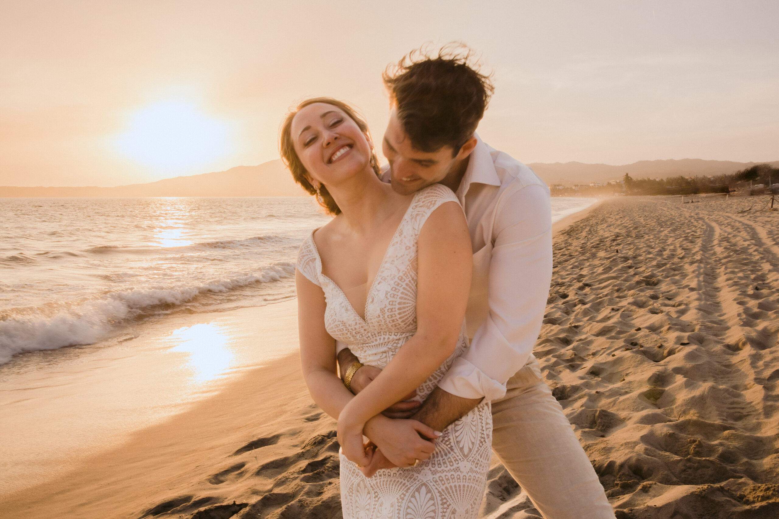 groom kissing bride's neck while on beach at sunset.