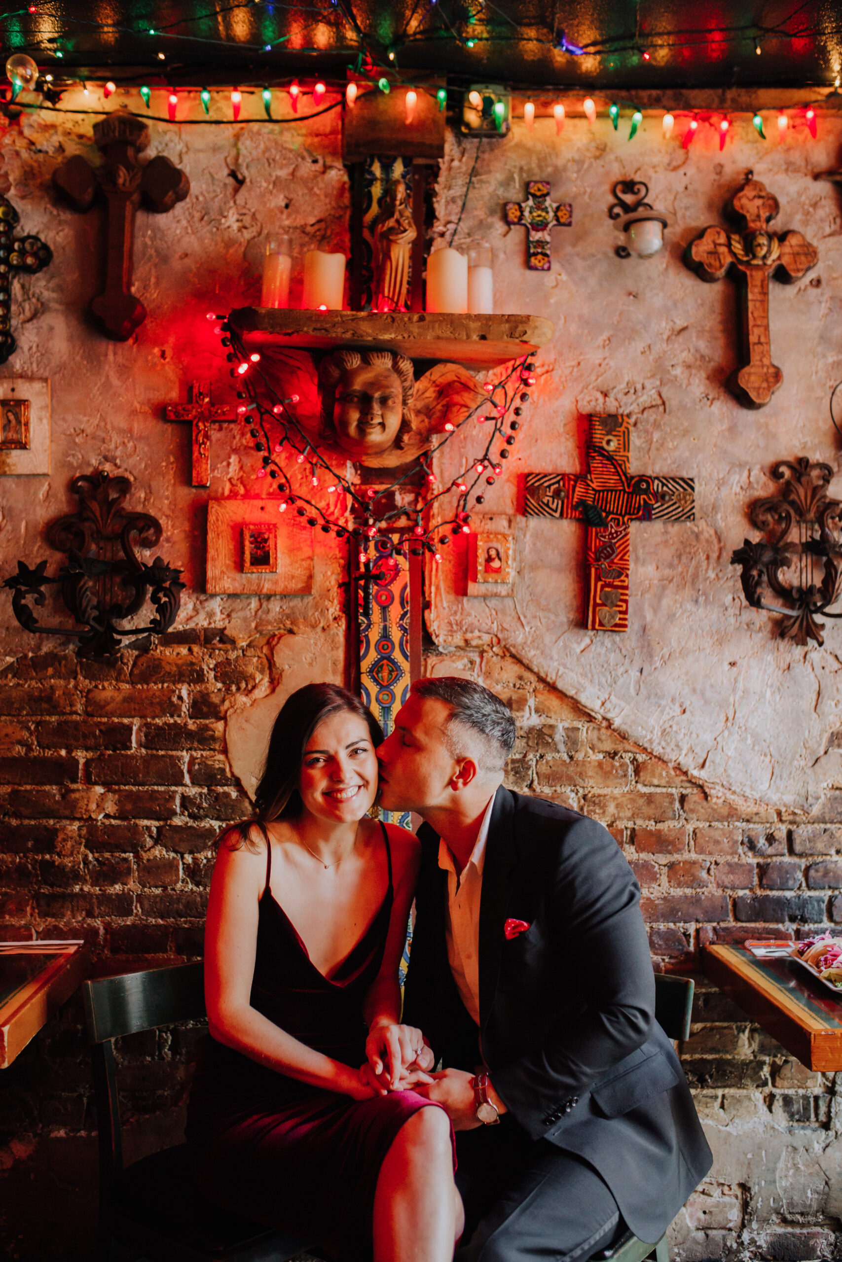 Beautiful couple sit in front of wall of crosses during their dreamy bar engagement photoshoot