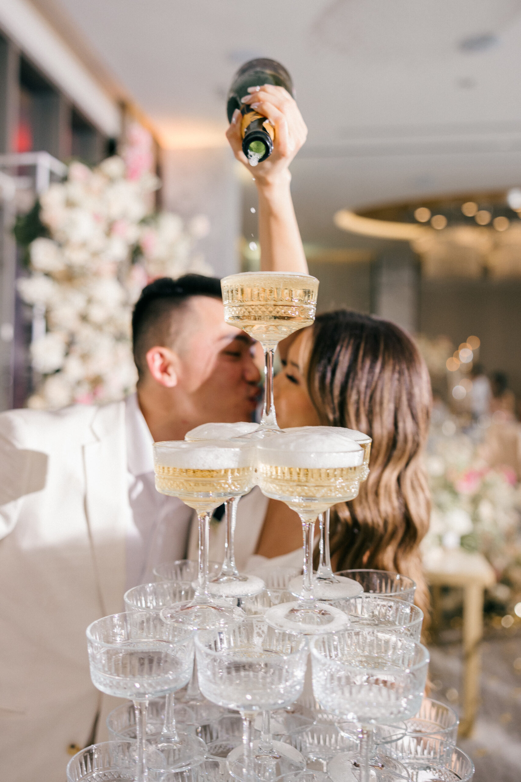 champagne tower gets filled with bride and groom kissing in the background